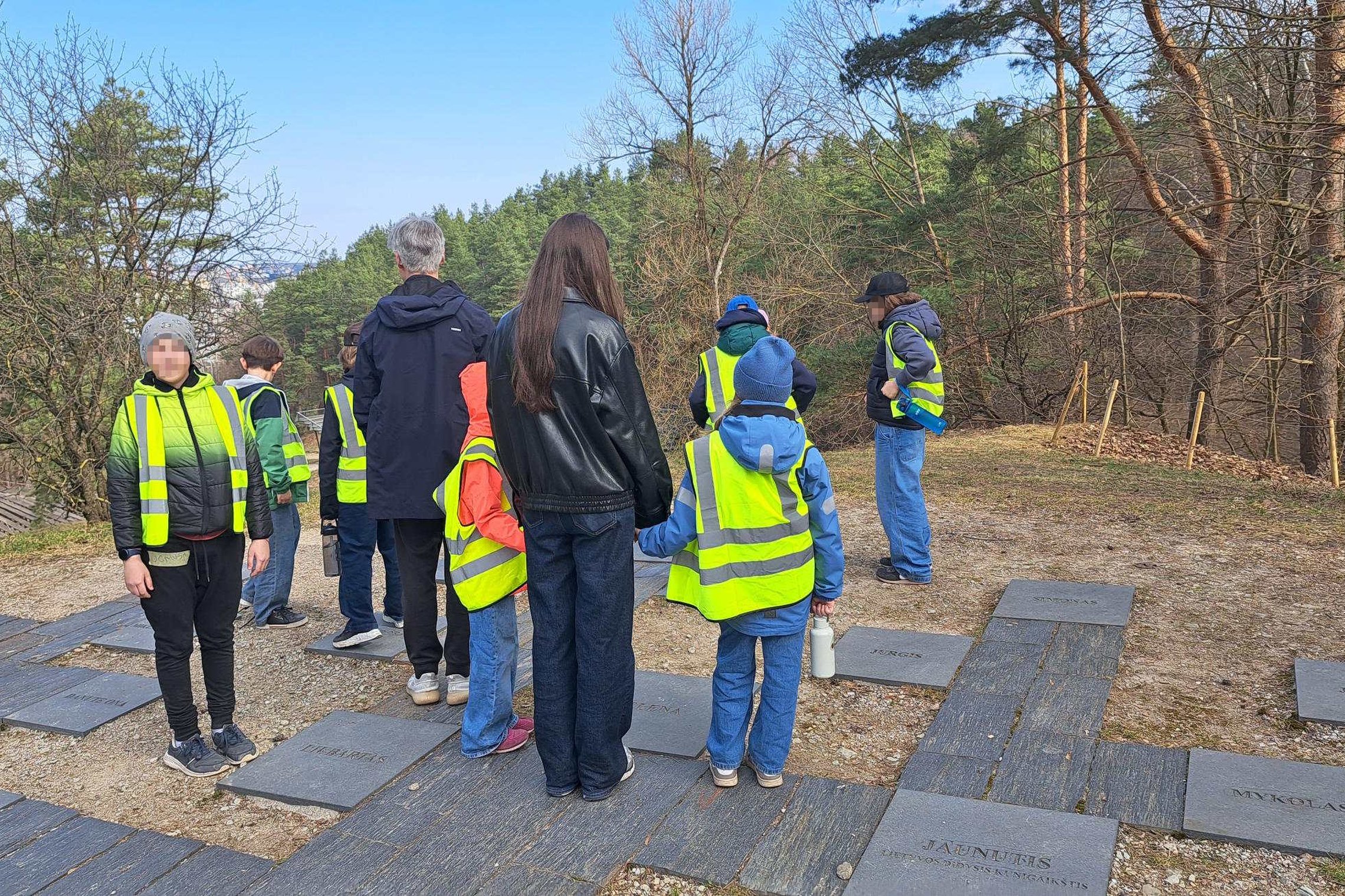 Grupė vaikų ir suaugusiųjų su ryškiomis saugos liemenėmis stovi Vilniaus pilių teritorija miško pakraštyje prie takelio su akmens plokštėmis, klausosi gido ir žvelgia į apylinkes nuo kalvos šlaito.