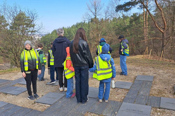 Grupė vaikų ir suaugusiųjų su ryškiomis saugos liemenėmis stovi Vilniaus pilių teritorija miško pakraštyje prie takelio su akmens plokštėmis, klausosi gido ir žvelgia į apylinkes nuo kalvos šlaito.
