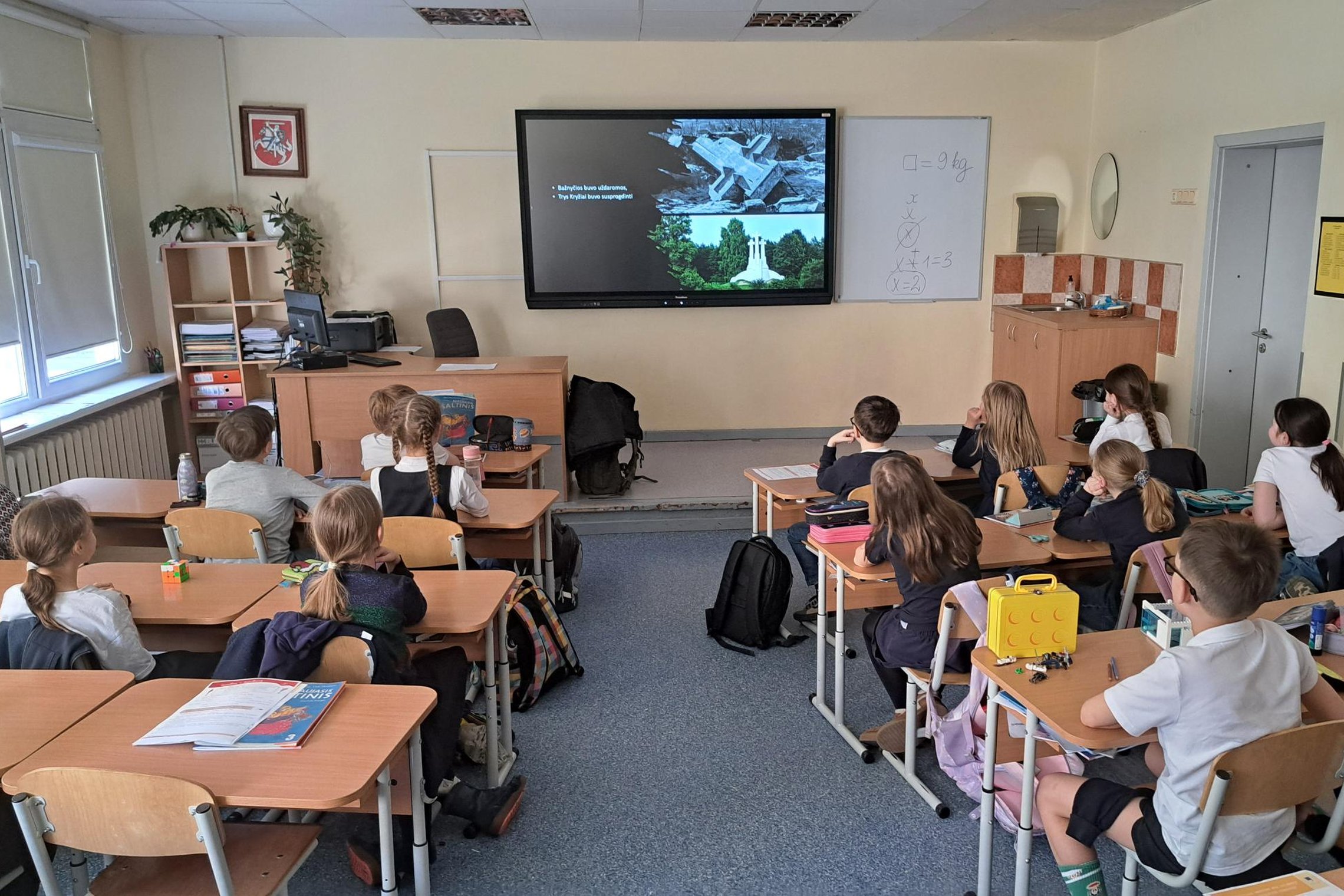 Trečiokai sėdi klasėje prie suolų ir žiūri į priekyje esančiame ekrane rodomą edukacinę skaidrę.
