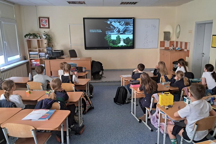 Trečiokai sėdi klasėje prie suolų ir žiūri į priekyje esančiame ekrane rodomą edukacinę skaidrę.