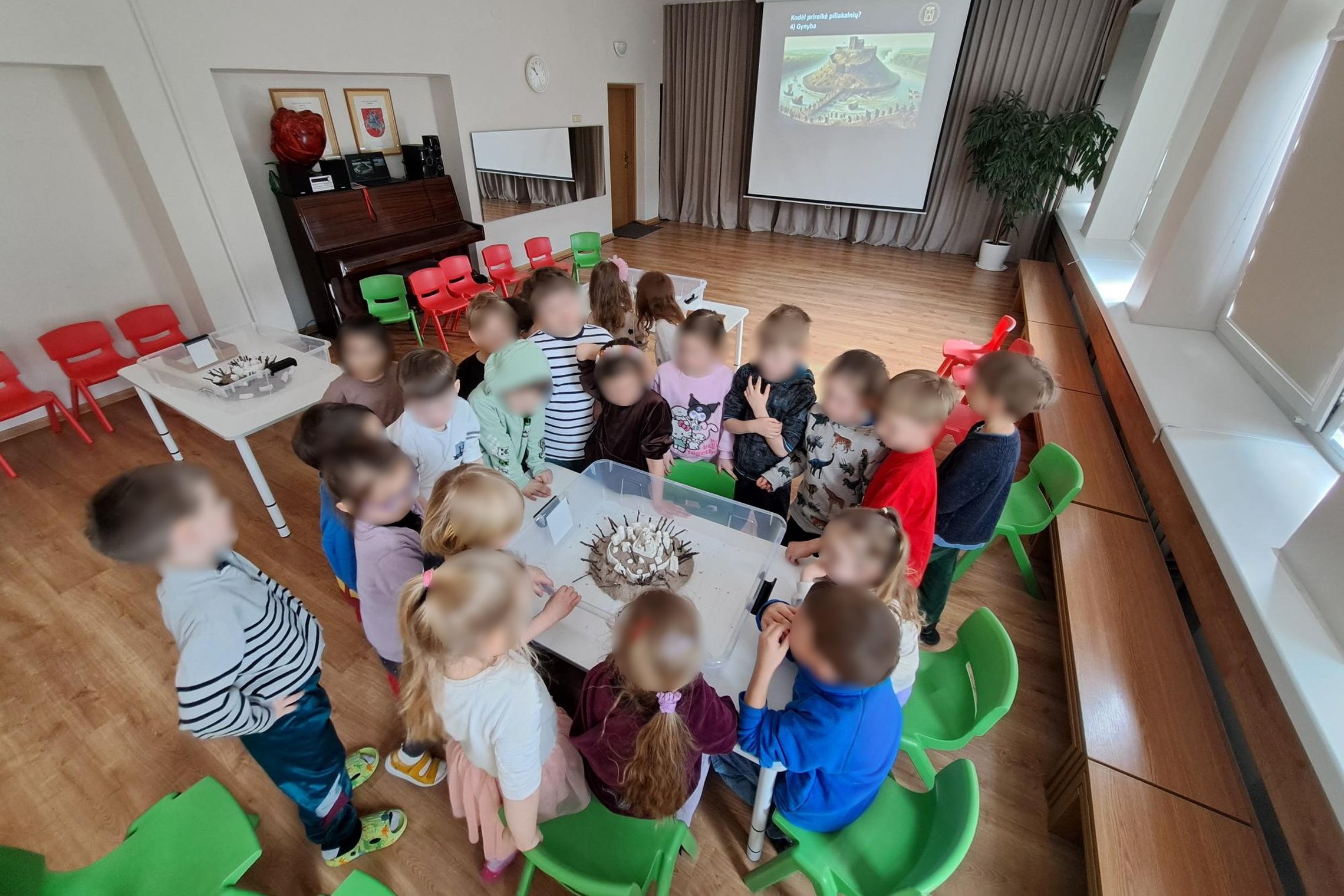 Nuotraukoje darželio aktų salė. Joje prie vieno stalo susibūrę vaikai. Ant stalo dėžė, joje piliakalnio modelis iš smėlio ir pilis viršuje. Šį piliakalnio modelį supylė ir pilį įtvirtino vaikai.