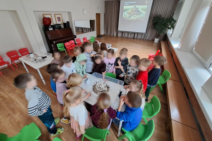 Nuotraukoje darželio aktų salė. Joje prie vieno stalo susibūrę vaikai. Ant stalo dėžė, joje piliakalnio modelis iš smėlio ir pilis viršuje. Šį piliakalnio modelį supylė ir pilį įtvirtino vaikai.