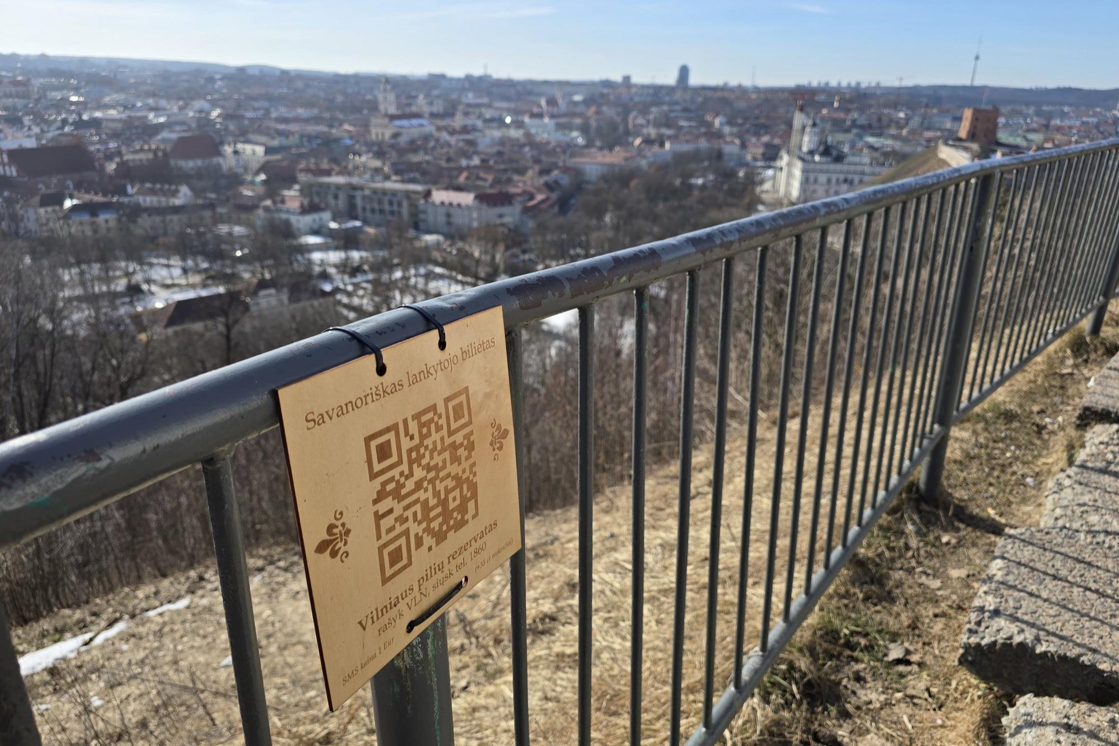 QR kodas su užrašu „Savanoriškas lankytojo bilietas“ pritvirtintas prie turėklo, fone matoma Vilniaus panorama nuo kalno.
