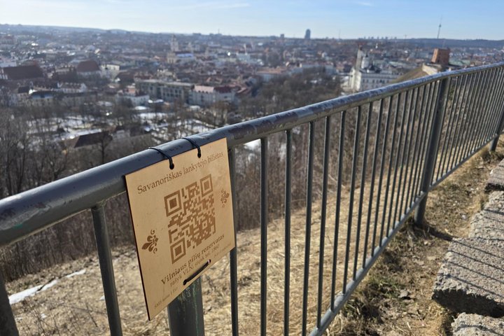QR kodas su užrašu „Savanoriškas lankytojo bilietas“ pritvirtintas prie turėklo, fone matoma Vilniaus panorama nuo kalno.