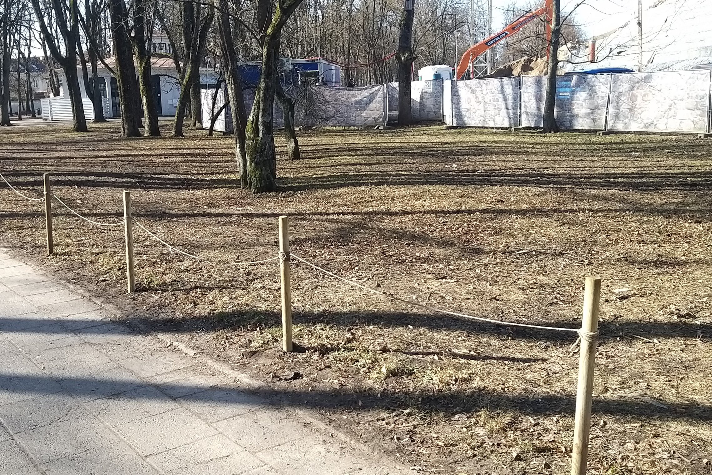 Pėsčiųjų takas palei mediniais kuoliukais ir virve pažymėtą želdyno kraštą miesto parke; tolumoje matyti medžiai ir statybų aptvėrimas.