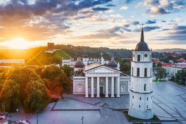 Vilniaus pilių teritorija saulėtekio metu: pirmame plane stovi Vilniaus katedra su varpine, aikštė beveik tuščia. Už medžių kyla Gedimino kalnas su Aukštutinės pilies bokštu, apšviestu tekančios saulės spindulių. Dangus nuspalvintas oranžiniais ir mėlynai