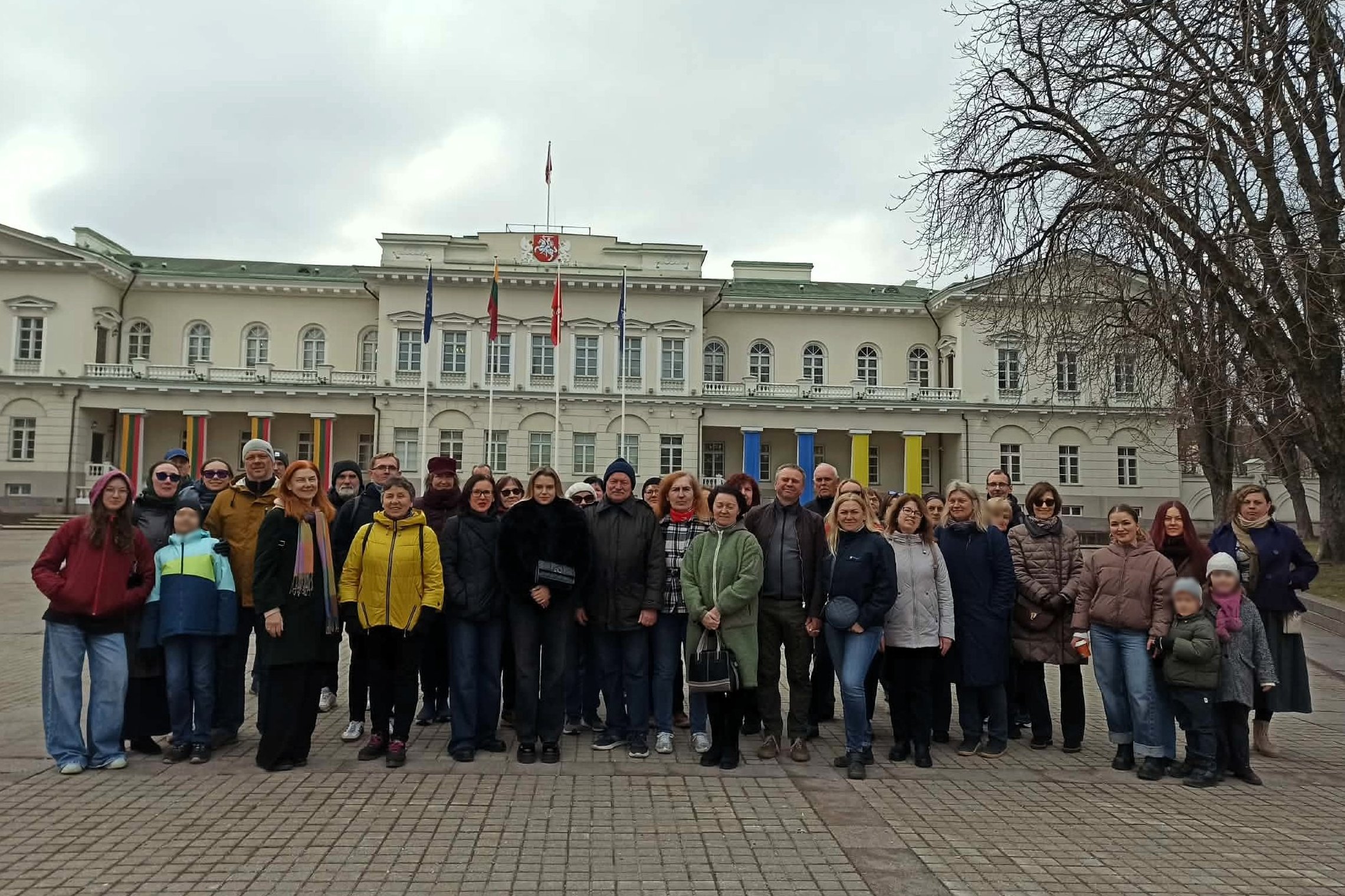 Didelė žmonių grupė stovi Vilniaus Prezidentūros kieme priešais šviesų istorinį pastatą su kolonomis ir Lietuvos vėliavomis. Dalyviai apsirengę šiltais drabužiais, pozuoja bendrai nuotraukai debesuotą dieną.