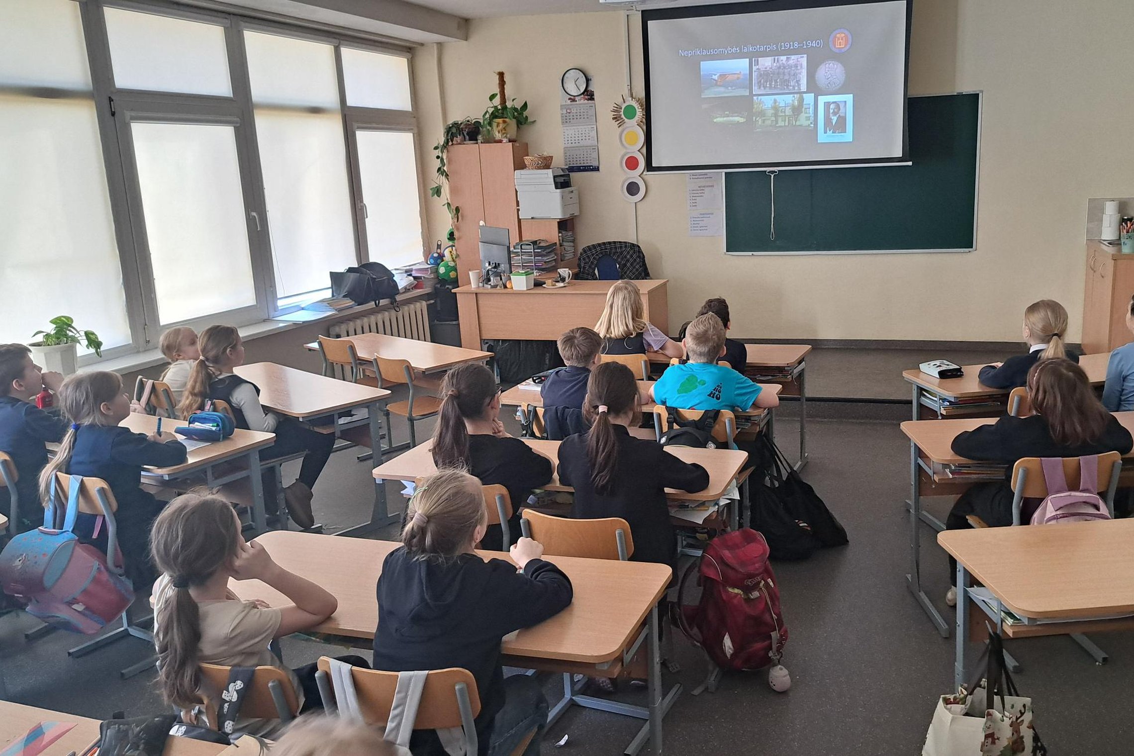 Trečiokai klasėje sėdi suoluose ir klausosi pristatymo apie Lietuvos nepriklausomybės laikotarpį, rodomo projektoriumi ekrane priešais lentą.