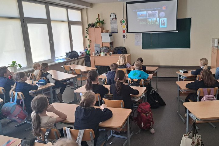 Trečiokai klasėje sėdi suoluose ir klausosi pristatymo apie Lietuvos nepriklausomybės laikotarpį, rodomo projektoriumi ekrane priešais lentą.