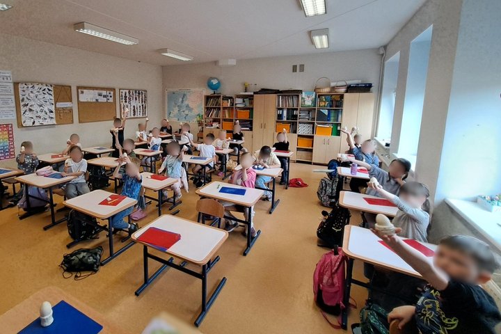 Nuotraukoje mokyklos klasė pilna suolų. Suoluose sėdi mokiniai. Mokiniai rankose laiko savo sukurtus margučių "sostus".
