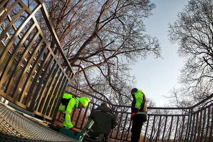 Trys darbininkai su ryškiomis darbo striukėmis remontuoja Altanos bokštelio terasos turėklą, saulėtą dieną tarp medžių.