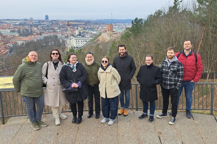 RIMI darbuotojų grupė stovi ant apžvalgos aikštelės Vilniaus pilių teritorijoje, fone matyti Gedimino pilies bokštas ir Vilniaus senamiesčio panorama.