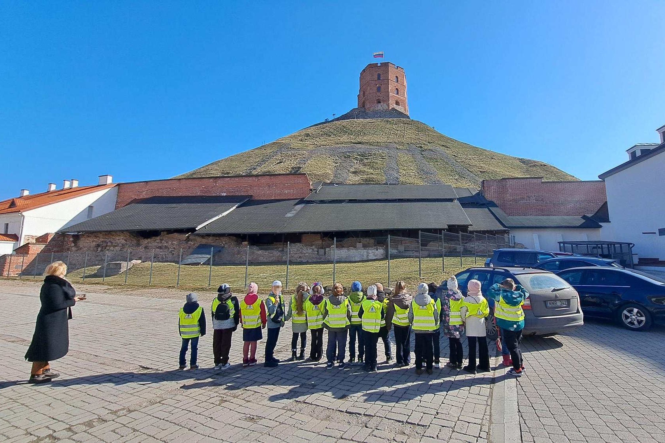 Pirmokų grupė su ryškiomis saugos liemenėmis Vilniaus pilių teritorijoje klausosi edukatorės ir žiūri į Gedimino pilies bokštą ant Pilies kalno.