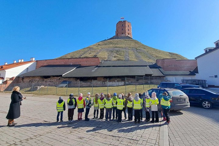 Pirmokų grupė su ryškiomis saugos liemenėmis Vilniaus pilių teritorijoje klausosi edukatorės ir žiūri į Gedimino pilies bokštą ant Pilies kalno.