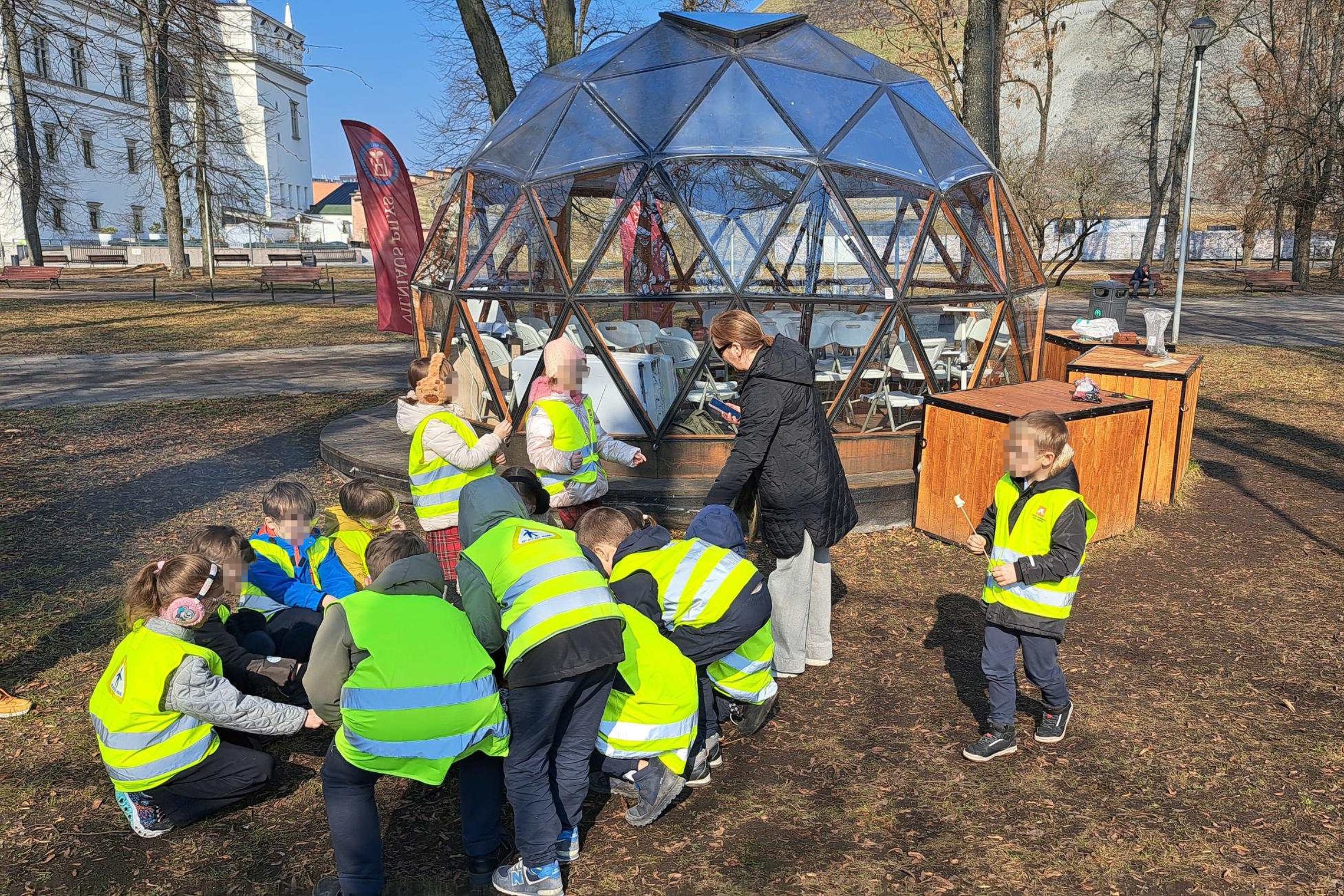 Vaikai su ryškiomis saugos liemenėmis Vilniaus pilių teritorijoje dalyvauja edukacijoje apie ugnį prie kupolo formos paviljono.