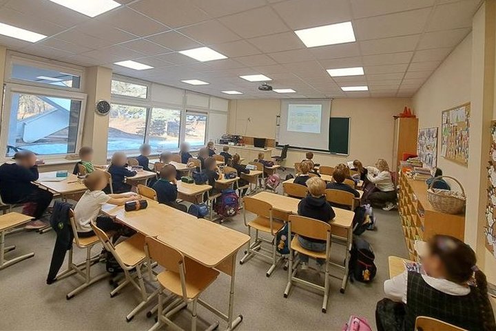 Nuotraukoje klasė. Šviesi, didelė ir pilna vaikų. Jie sėdi suoluose ir žiūri į ekraną. Nuotrauka daryta vaikams iš nugarų, veidų nesimato. Bet ekrane puikiai matyti edukacijos tema.