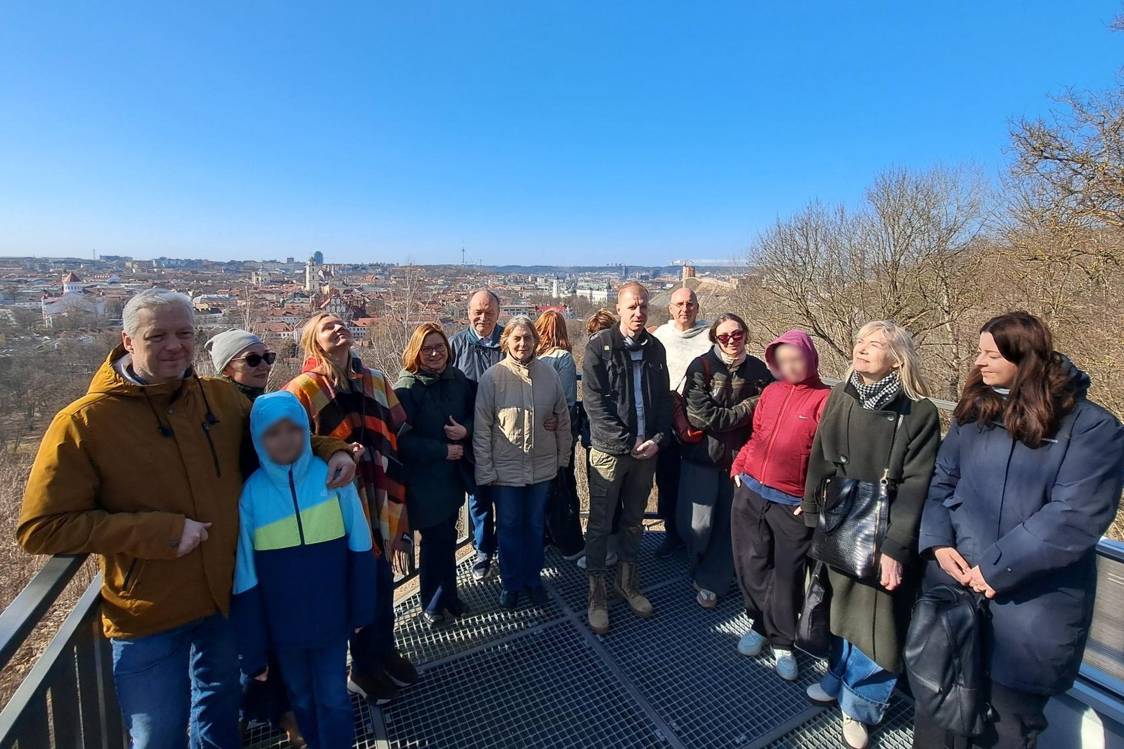 Nuotraukoje grupė žmonių stovi Altanos bokštelio terasoje. Už jų atsiveria viena gražiausių Vilniaus panoramų – senamiesčio bokštai, stogai ir miesto horizontas.