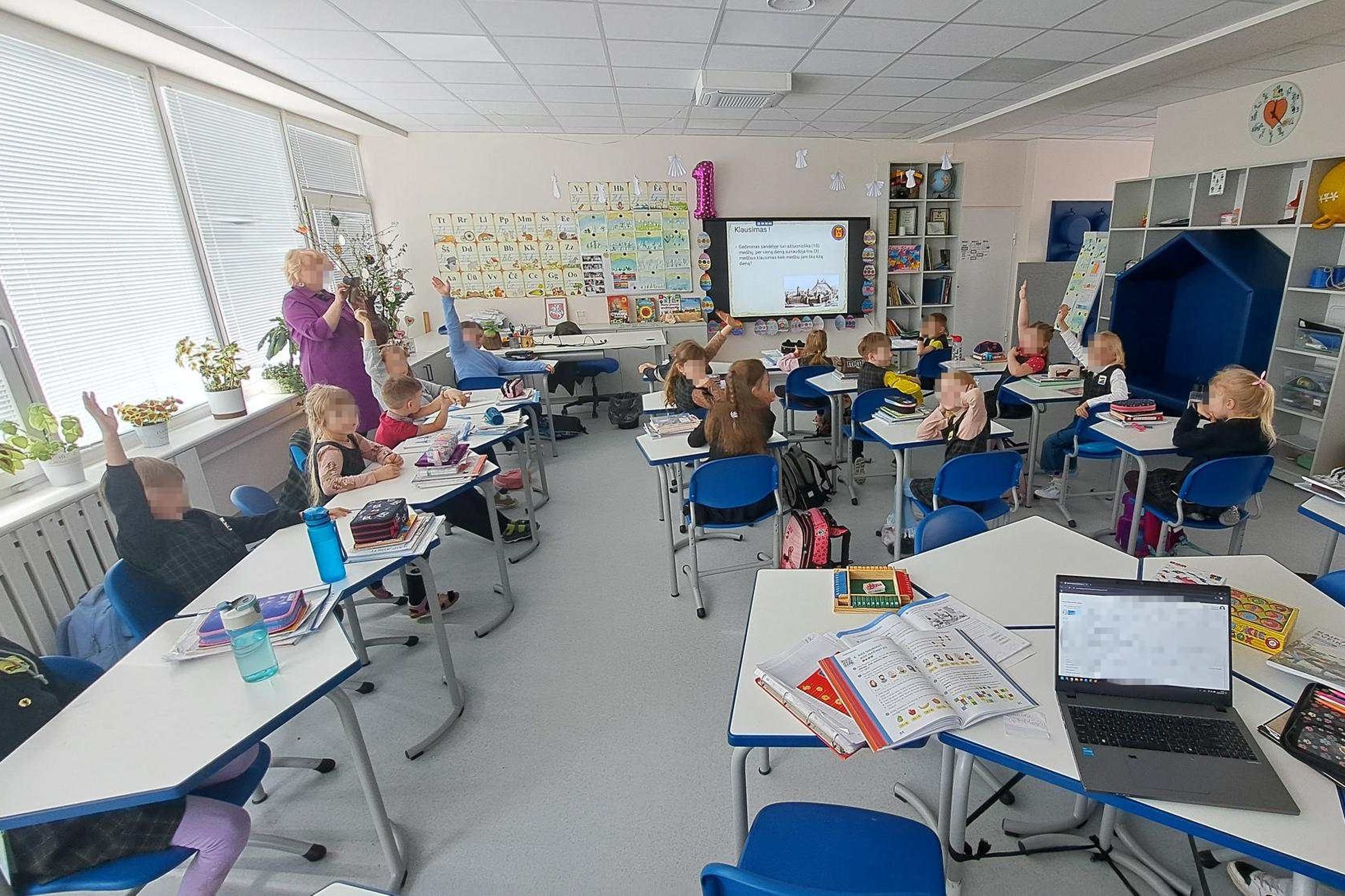 Pirmokai klasėje kelia rankas ir dalyvauja edukacijoje, priekyje rodomos skaidrės.