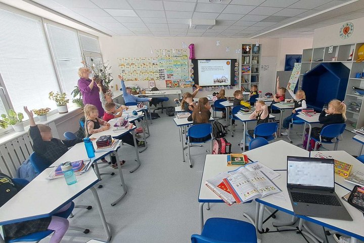 Pirmokai klasėje kelia rankas ir dalyvauja edukacijoje, priekyje rodomos skaidrės.