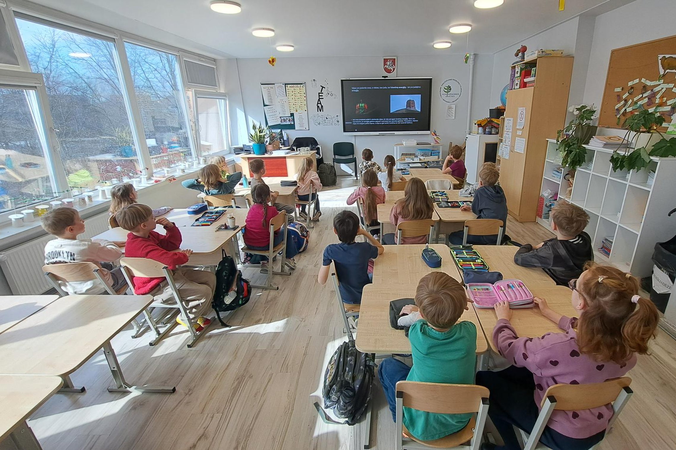 Klasėje prie suolų sėdintys pradinukai stebi edukacinę medžiagą ekrane apie vėjo energiją.