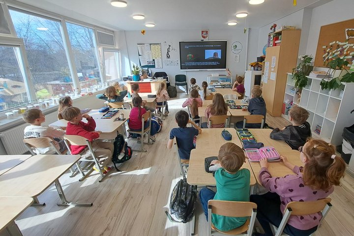 Klasėje prie suolų sėdintys pradinukai stebi edukacinę medžiagą ekrane apie vėjo energiją.