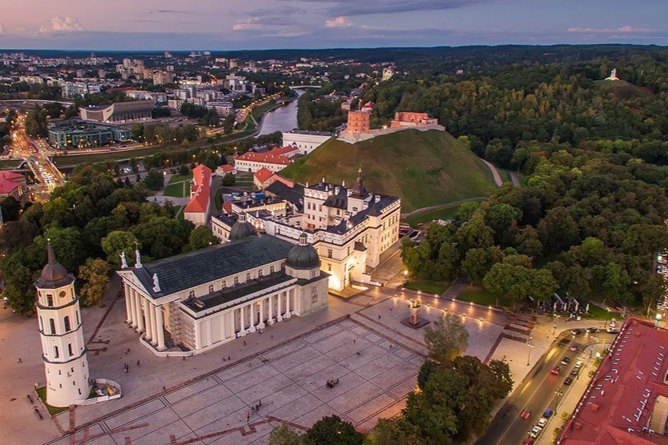 Vilniaus senamiesčio panorama iš paukščio skrydžio: priekyje – Katedros aikštė su balta Vilniaus katedra ir varpine, už jos – Gedimino pilies bokštas ant kalno, tolumoje vingiuoja Neris, o miestą supa žali parkai ir vakaro dangus.