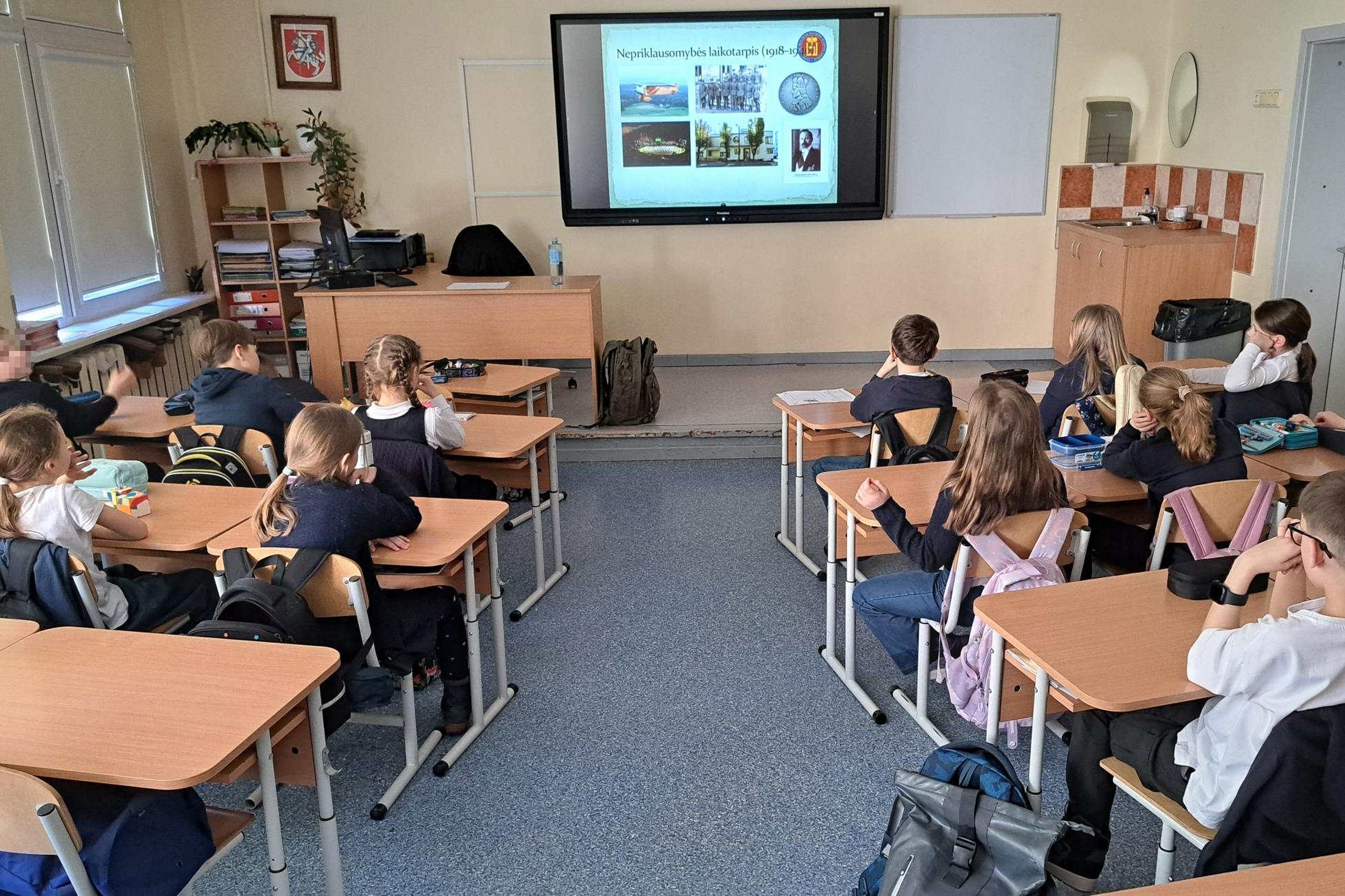 Trečiokų klasė sėdi mokyklos kabinete prie suolų ir klausosi edukacijos apie Lietuvos nepriklausomybės laikotarpį – priekyje matomas ekranas su skaidre „Nepriklausomybės laikotarpis “