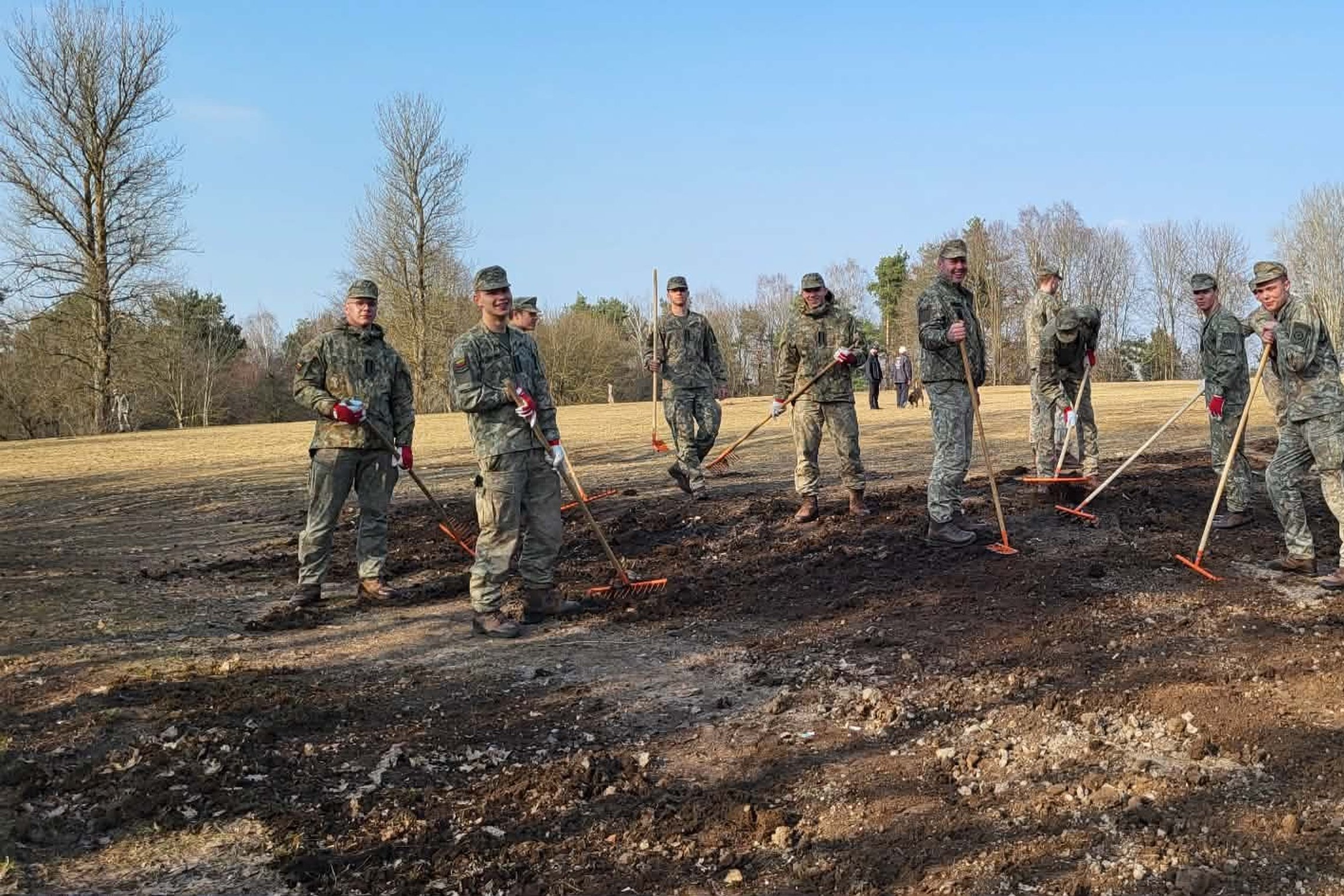 Grupė karių su uniformomis stovi lauke ir grėbliais tvarko sudegusios žemės plotą. Aplink matyti medžiai ir atvira pieva, diena saulėta.