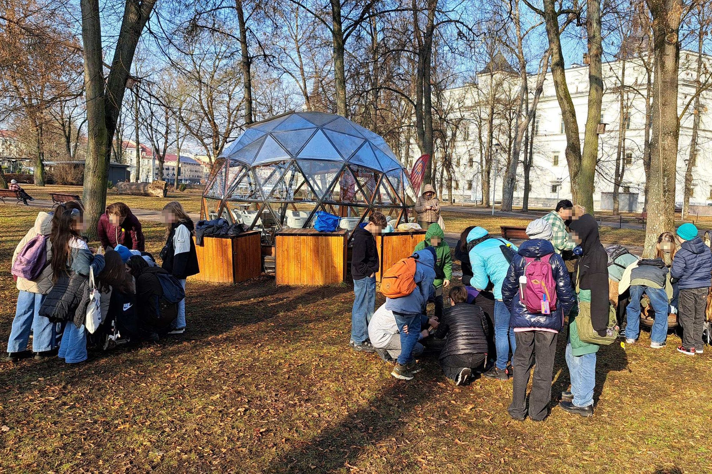 Moksleivių grupė dalyvauja edukacijoje Vilniaus pilių teritorijoje prie stiklinio kupolo, klausosi edukatoriaus ir atlieka praktinę užduotį ant žemės.