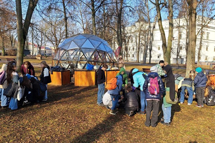 Moksleivių grupė dalyvauja edukacijoje Vilniaus pilių teritorijoje prie stiklinio kupolo, klausosi edukatoriaus ir atlieka praktinę užduotį ant žemės.