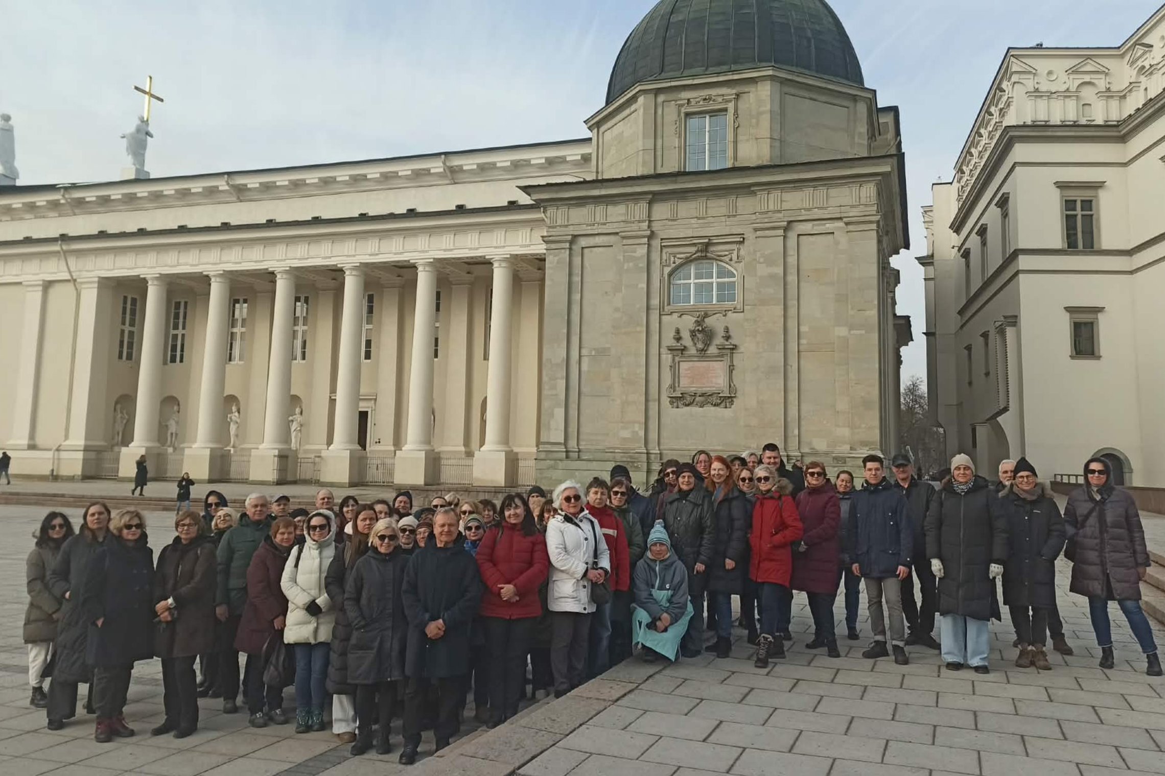Grupė žmonių stovi Vilniaus katedros aikštėje priešais Vilniaus arkikatedrą baziliką su kolonomis ir kupolu, fotografuojasi po ekskursijos vėsią ankstyvo pavasario dieną.