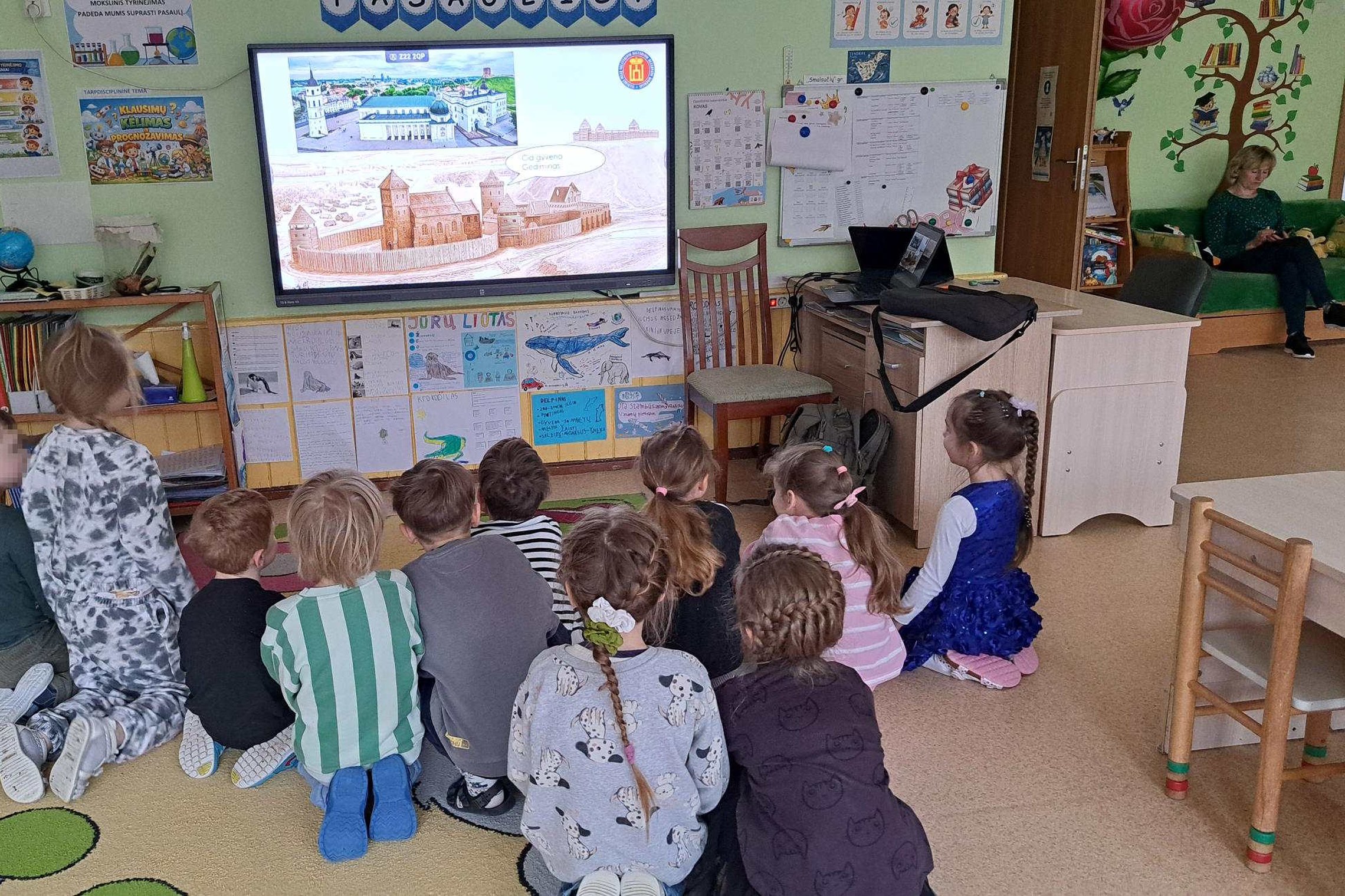 Darželinukai sėdi ant kilimo ir stebi edukaciją apie Gediminą ir Vilniaus pilis. Priekyje matomas ekranas su vaizdine mokymo medžiaga.