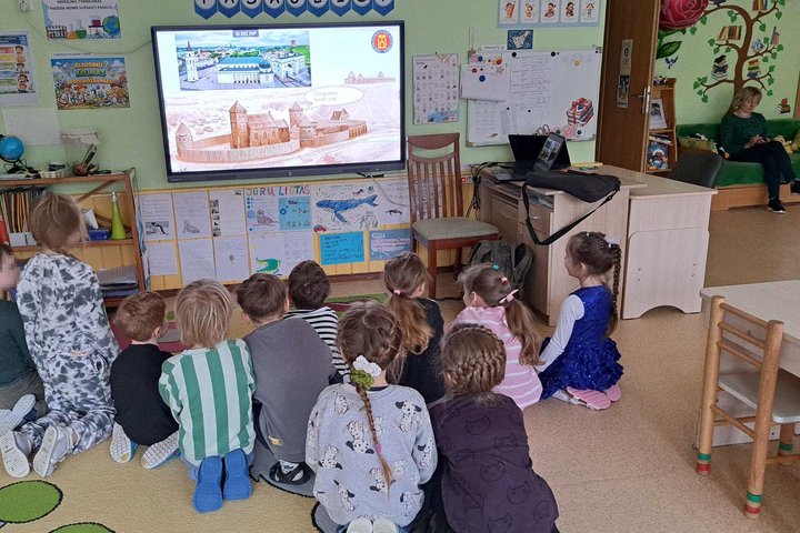 Darželinukai sėdi ant kilimo ir stebi edukaciją apie Gediminą ir Vilniaus pilis. Priekyje matomas ekranas su vaizdine mokymo medžiaga.