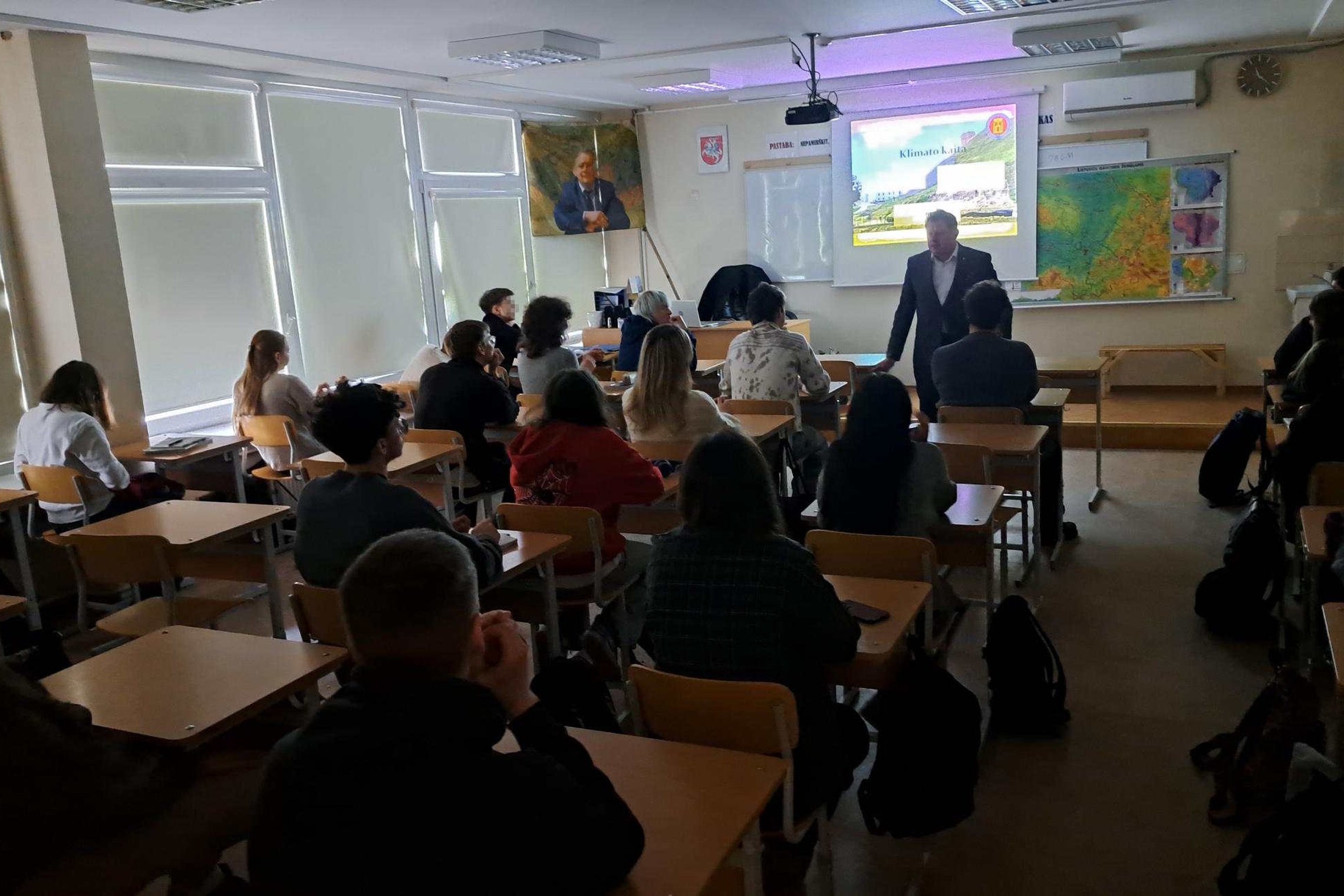 Mokyklos klasėje vyksta edukacija – mokiniai sėdi suoluose ir klausosi priekyje stovinčio lektoriaus, o ekrane rodomas pristatymas apie klimato kaitą; ant sienų matyti žemėlapiai, lenta ir portretas.