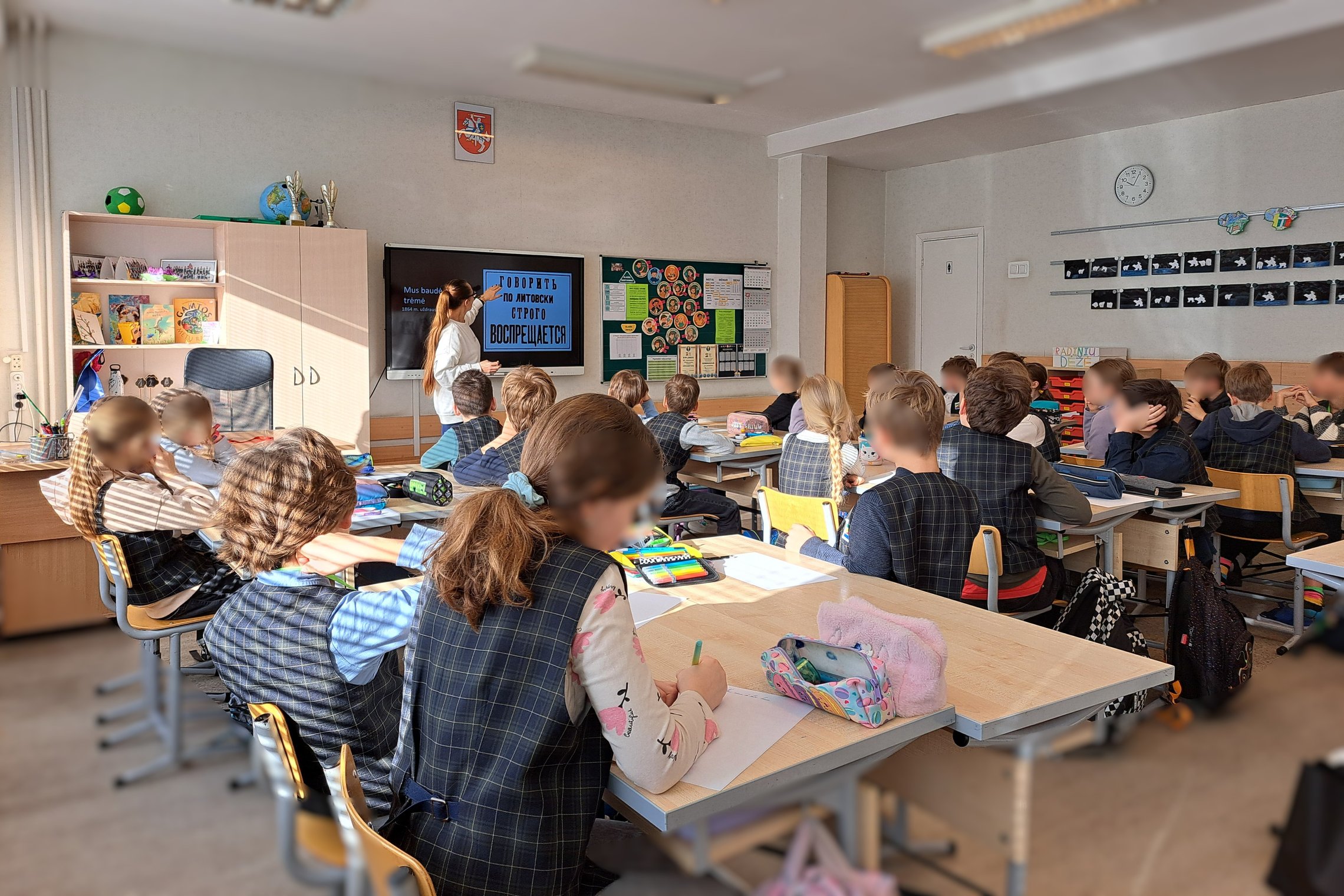 Nuotraukoje klasė pilna mokinių. Jie nusisukę žiūri į ekraną. Jame edukatorė rodo edukacijos medžiagą.