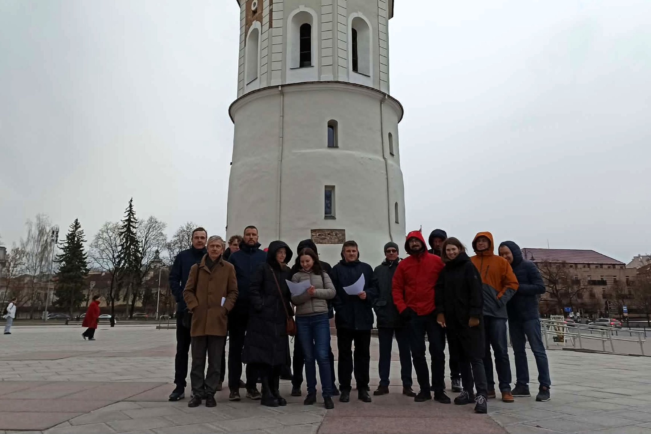 Ekskursijos dalyvių grupė stovi prie Gedimino bokšto Vilniaus pilių teritorijoje, laikydami lapus rankose.