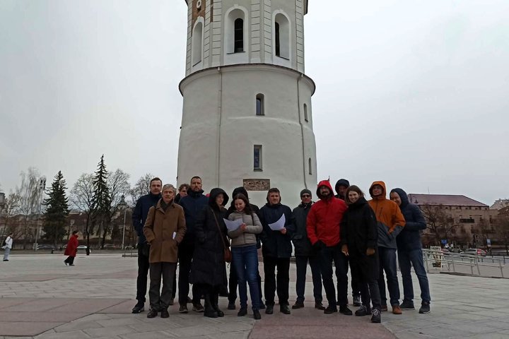 Ekskursijos dalyvių grupė stovi prie Gedimino bokšto Vilniaus pilių teritorijoje, laikydami lapus rankose.