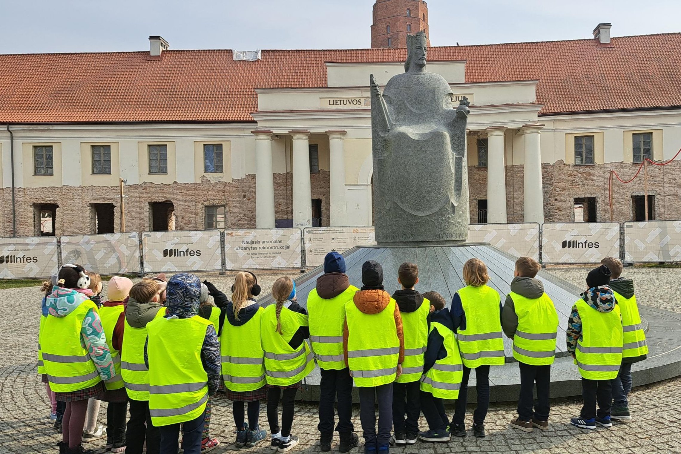 Antrokai su ryškiomis geltonomis liemenėmis stovi Vilniaus pilių teritorijoje ir klausosi pasakojimo prie Mindaugo paminklo, fone matyti rekonstruojamas pastatas.