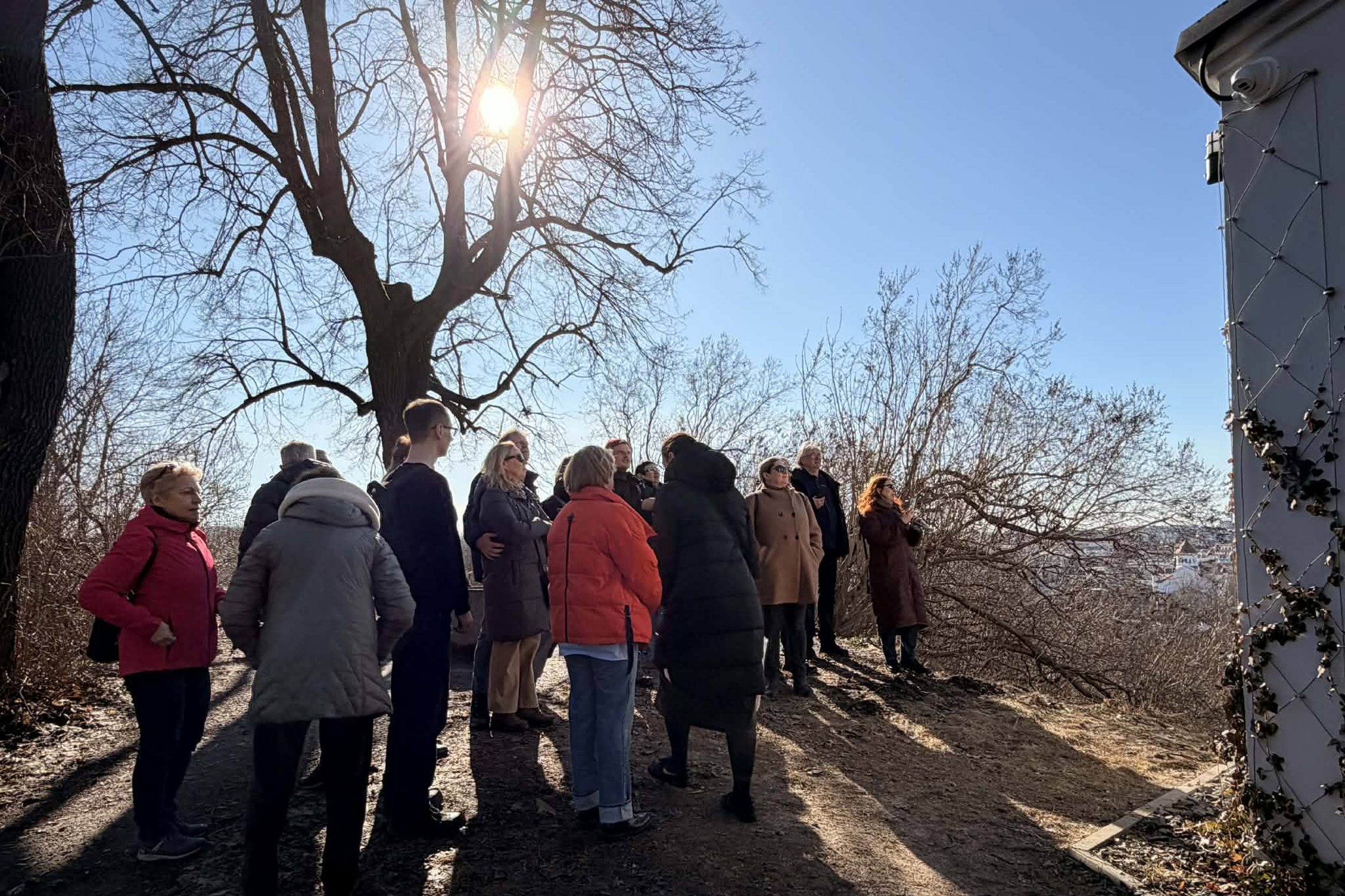 Ekskursijos dalyvių grupė stovi ant kalvos ir klausosi gido, saulėtą dieną žvelgdami į Vilniaus panoramą šalia pastato sienos.