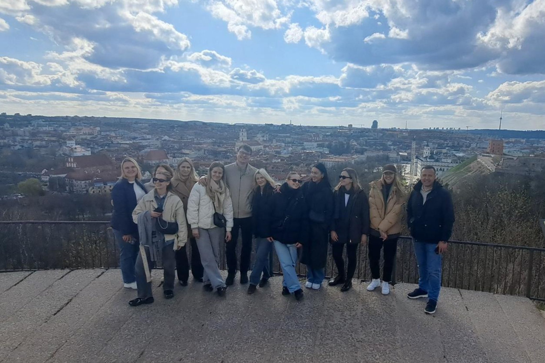 Grupė suaugusiųjų stovi ant apžvalgos aikštelės, fone atsiveria plati Vilniaus miesto panorama su senamiesčiu, kalvomis ir debesuotu dangumi.