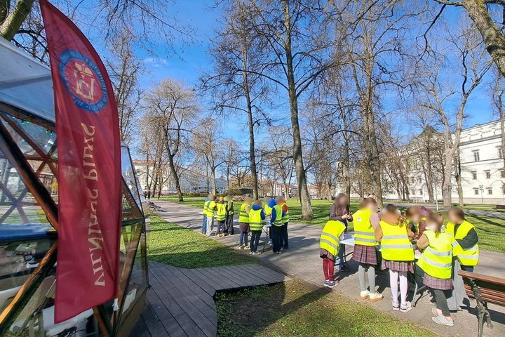 Vaikai su ryškiomis liemenėmis dalyvauja edukacijoje Vilniaus pilių kupolo teritorijoje, saulėtą dieną klausosi ir atlieka užduotis parke šalia medžių ir istorinių pastatų.