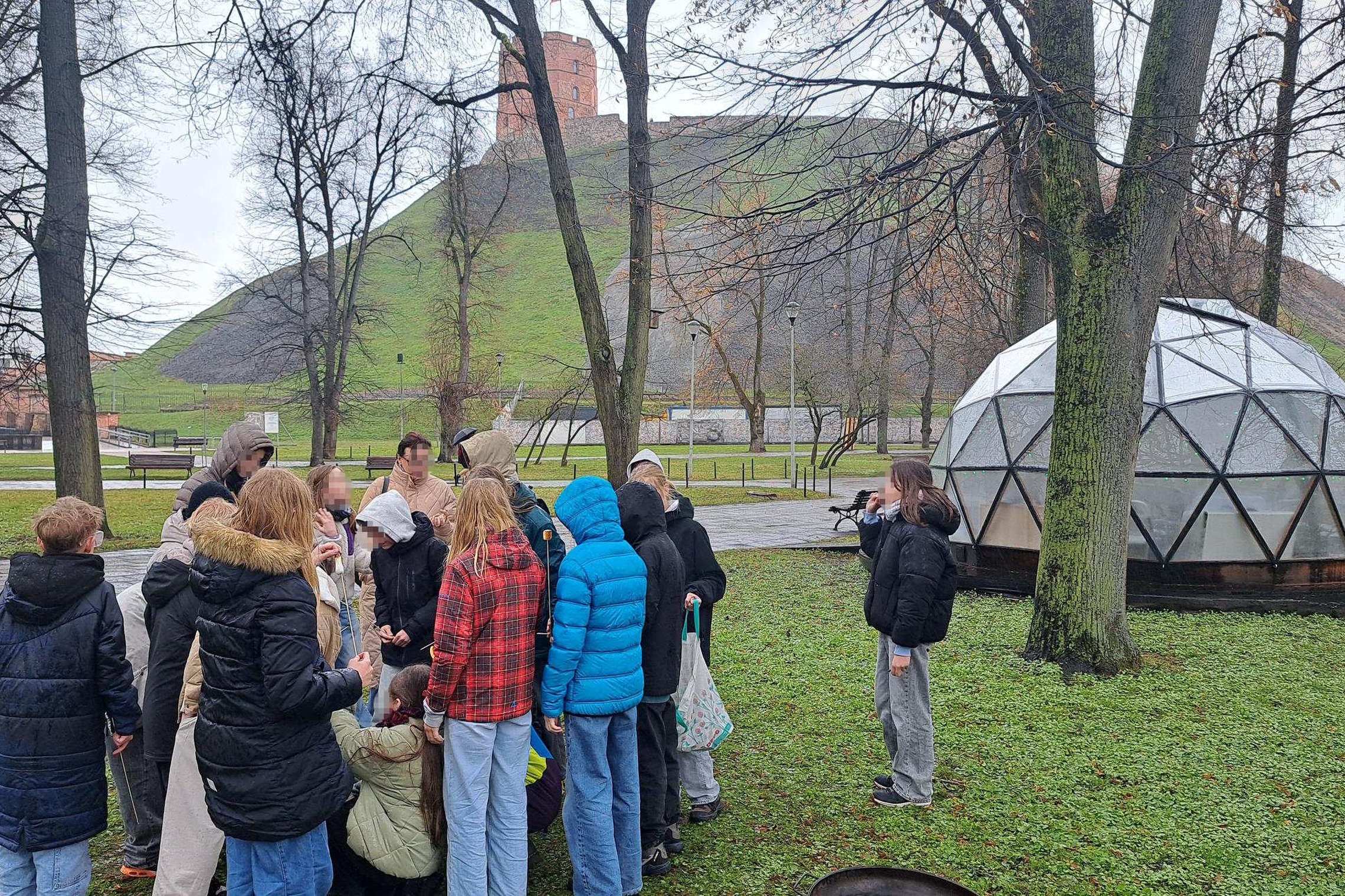 Grupė vaikų dalyvauja edukacijoje lauke prie Vilniaus pilių kupolo, fone matyti Gedimino pilies kalnas, o dalyviai susibūrę aplink vietą, kur mokosi kurti ugnį.