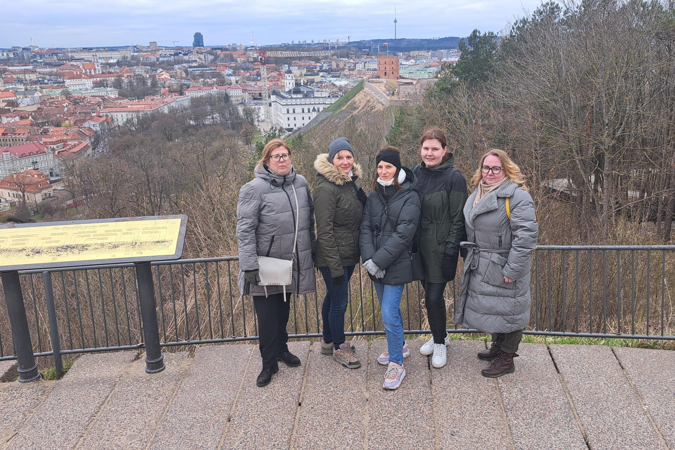 Penkių žmonių grupė stovi ant apžvalgos aikštelės Vilniaus pilių teritorijoje, fone matyti Gedimino bokštas ir miesto panorama debesuotą dieną.