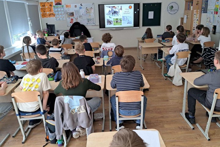 Mokiniai sėdi klasėje prie suolų ir klausosi edukacinės pamokos, kuri rodoma interaktyviame ekrane priekyje.