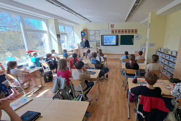 Klasėje prie suolų sėdi pradinių klasių mokiniai ir stebi priekyje rodomą pristatymą ekrane, pro langus sklinda saulės šviesa.