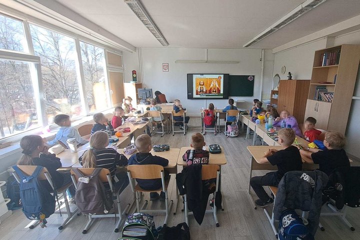 Pradinės klasės mokiniai sėdi klasėje prie suolų ir klausosi edukacijos, rodomos ekrane prie lentos, šviesioje patalpoje su dideliais langais ir baldais, tarp jų – spintos bei elektros skydinė.
