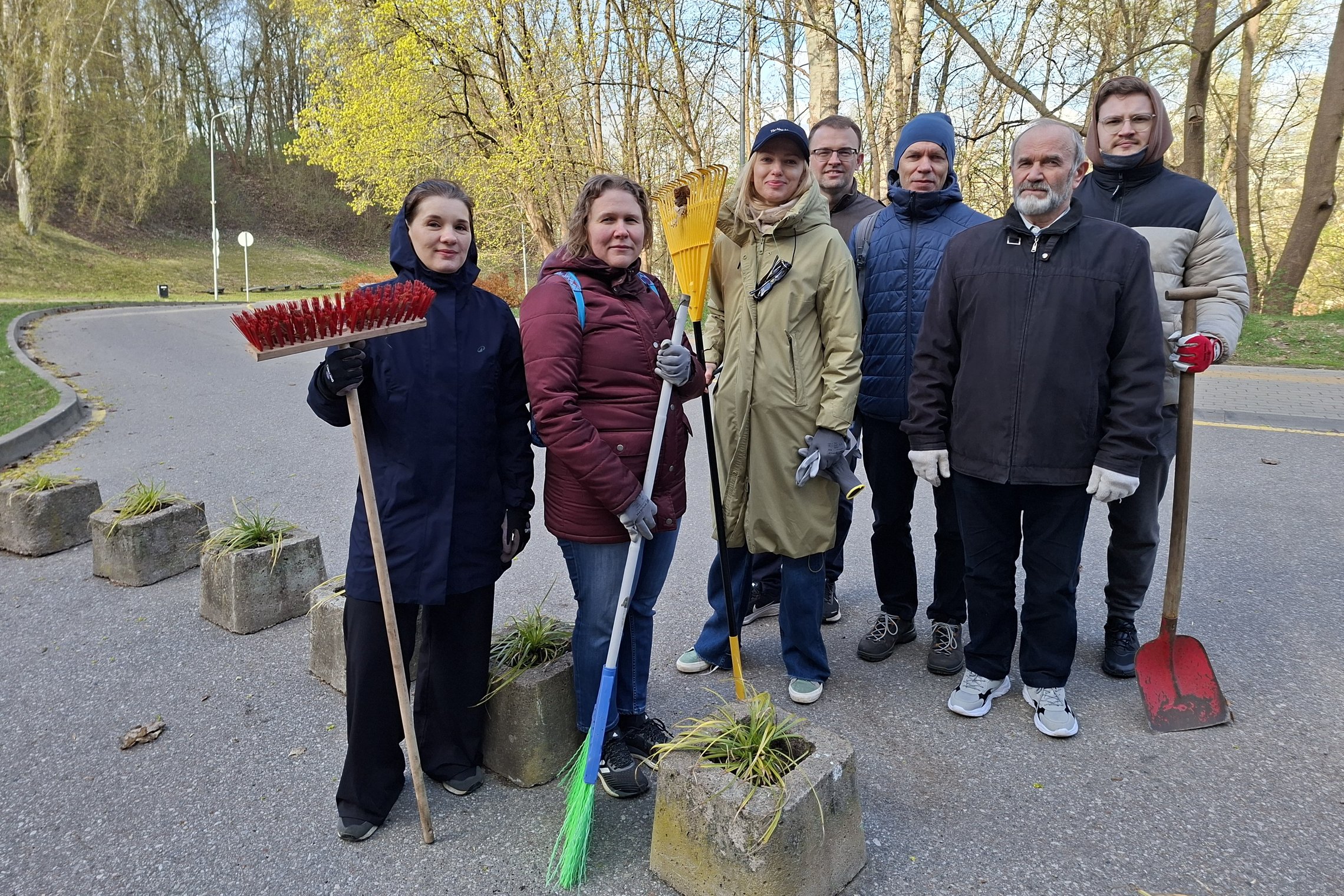 Grupė savanorių Vilniaus pilių teritorijoje stovi lauke su darbo įrankiais – grėbliais, šluotomis ir kastuvu. Jie apsirengę šiltais drabužiais, nusiteikę darbui. Aplink matyti takas, medžiai ir pavasariškai bundanti gamta.