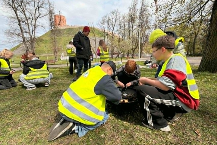 Mokiniai su ryškiaspalvėmis liemenėmis dalyvauja edukacinėje veikloje Vilniaus pilių teritorijoje, Gedimino kalno papėdėje. Susiskirstę į grupes jie atlieka praktines užduotis ant žolės, o fone matyti Gedimino kalnas ir Aukštutinės pilies bokšto liekanos.