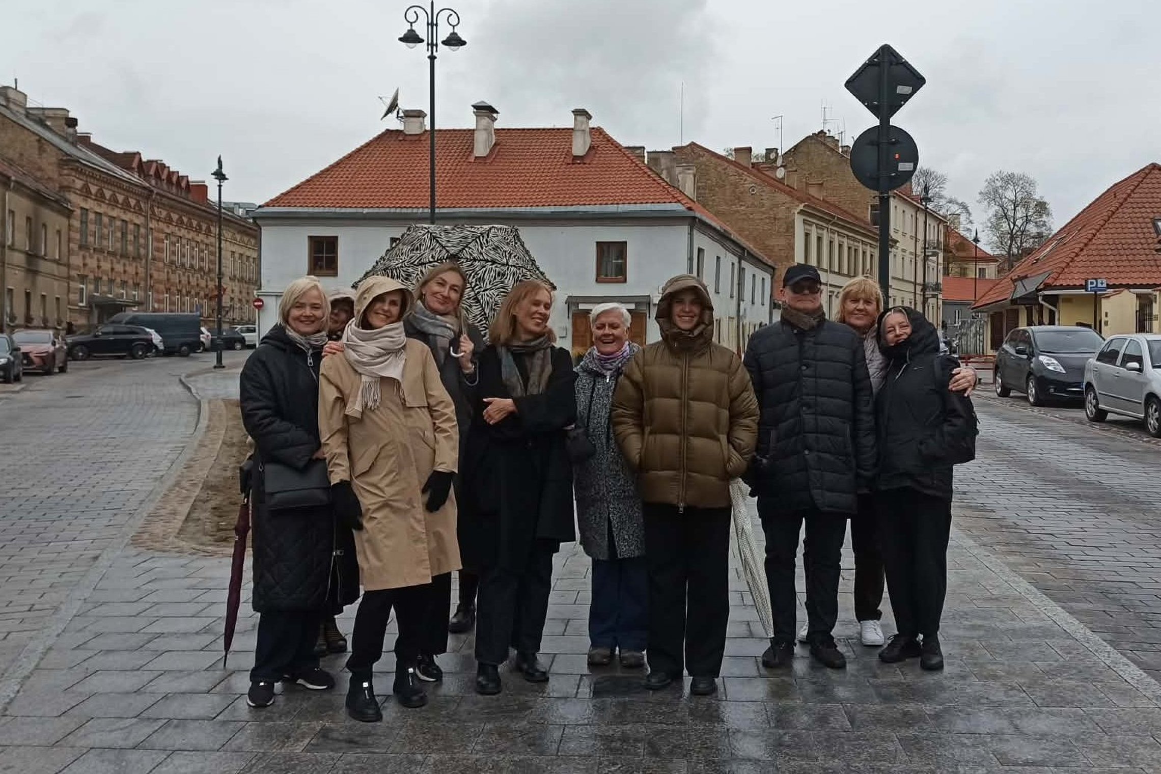 Grupė žmonių stovi Vilniaus senamiestyje lietingą dieną – apsirengę šiltais drabužiais, kai kurie su gobtuvais ar skėčiu. Jie šypsosi ir pozuoja nuotraukai ant šlapios grindinio gatvės, o už jų matyti istoriniai pastatai su raudonais stogais.