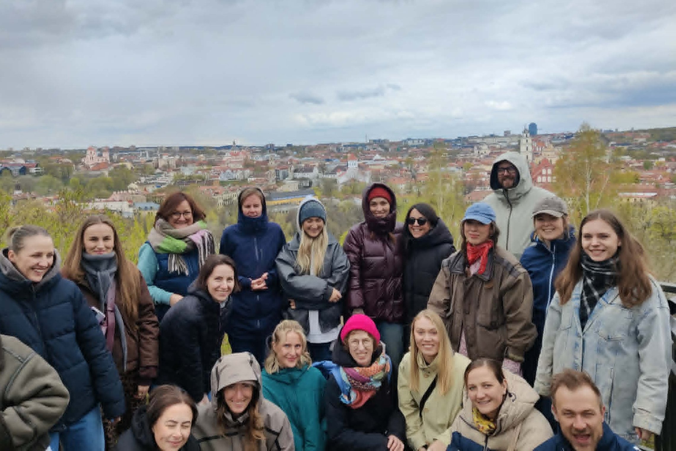 Grupinė ekskursijos dalyvių nuotrauka Vilniaus pilių teritorijoje, nuo kalvos atsiveriant miesto panoramai. Dalyviai apsirengę šiltais lauko drabužiais, šypsosi, o fone matyti Vilniaus senamiesčio stogai, bažnyčių bokštai ir pavasariškai žaliuojantys medž